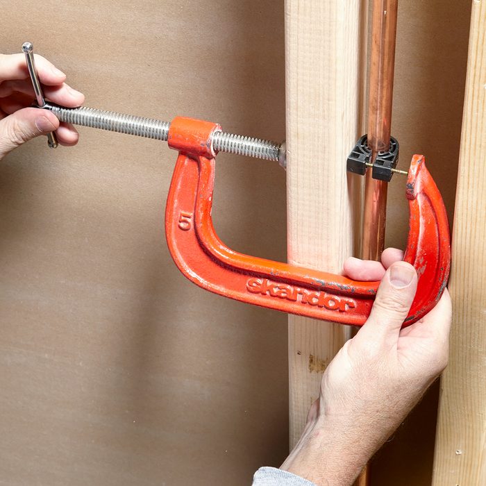 Hands using an orange C-clamp to hold copper piping against a wooden beam. The word "Sharidan" is visible on the clamp.