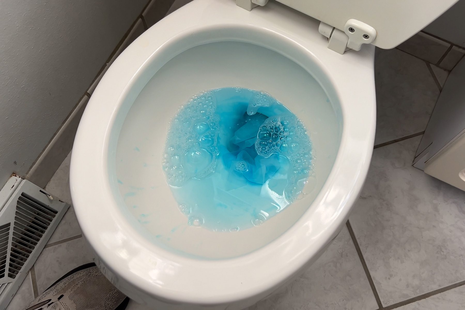 A close-up view of a toilet with blue cleaning solution in the water. The toilet is white, and the floor is made of light-colored tiles. A white heater is partially visible on the left side.