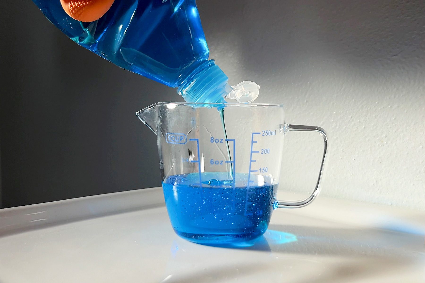 A bottle of blue liquid is being poured into a clear measuring pitcher, sitting on a white surface. The pitcher has measurement markings on it, and the scene is lit by natural light from the left.