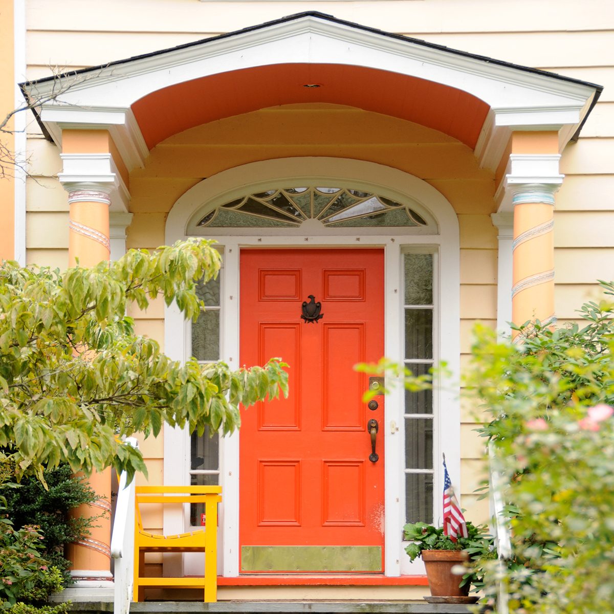 A vibrant orange front door with a decorative archway. The house exterior is pale yellow with white trim. A small American flag is placed in a pot with plants on the porch, and greenery surrounds the entrance.