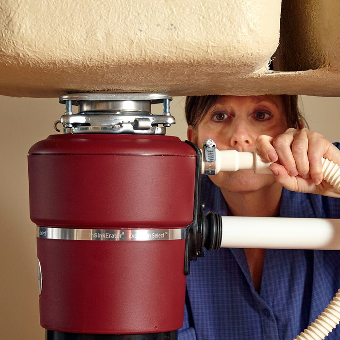 A person in a blue shirt inspects a red garbage disposal unit under a sink, adjusting connections. The focus is on the appliance and plumbing, highlighting a repair or installation process.