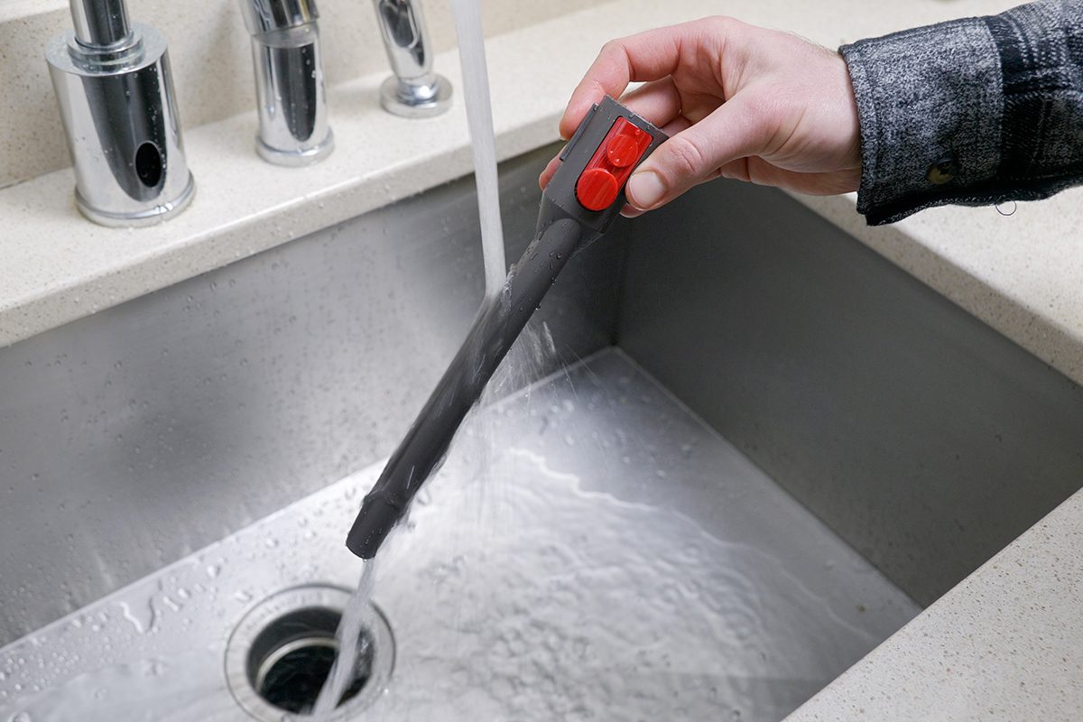 A person holding a grey faucet sprayer with red buttons, rinsing water into a stainless steel sink. The countertop is light-colored.
