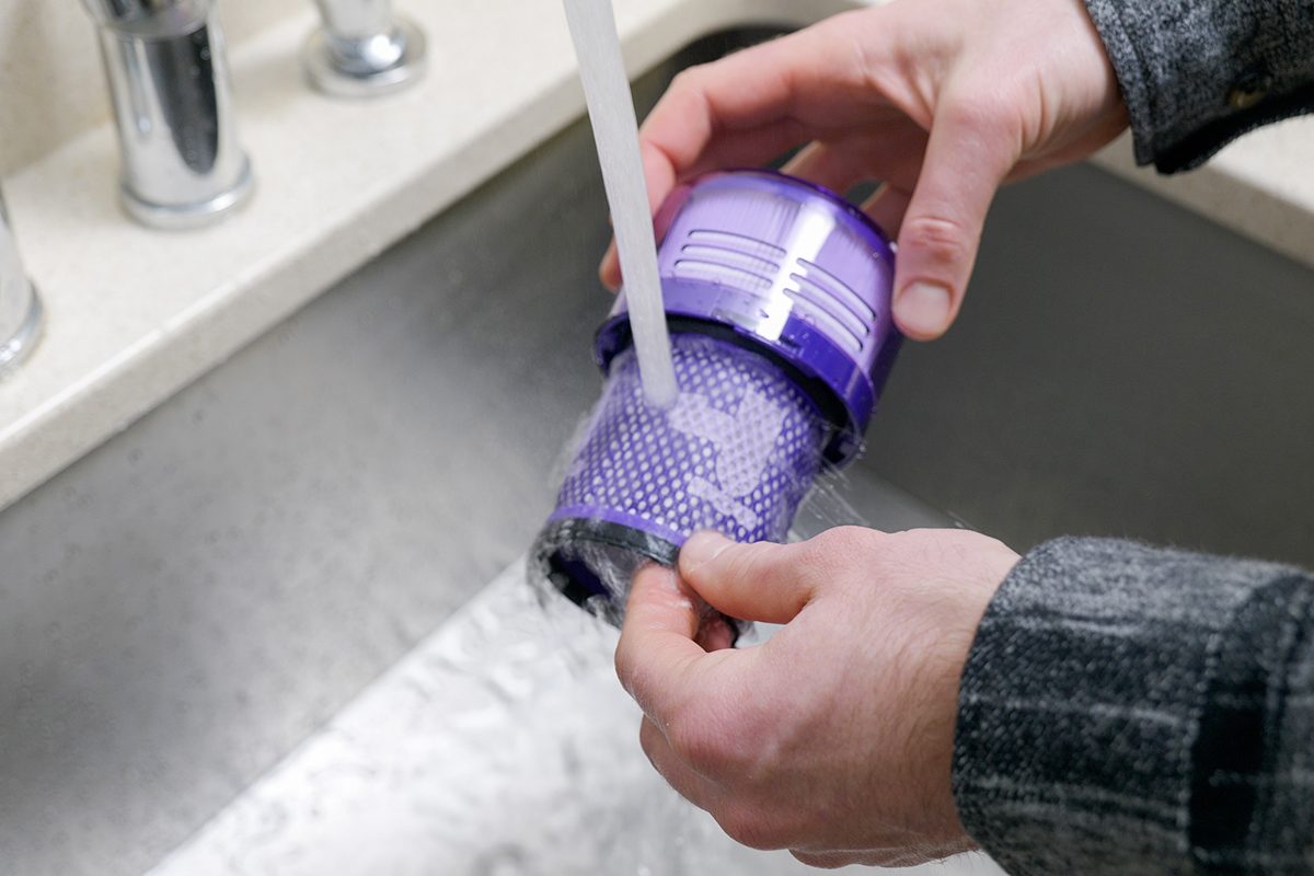 A person rinses a purple vacuum filter under a running tap in a sink. The person is wearing a dark plaid shirt. The water flows over the filter as it is being cleaned.