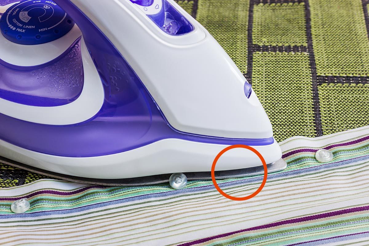 Purple and white iron with a groove between the plastic body and the metal iron plate, with a red circle around a section of the groove, on top of a green carpet and a multicolored striped button down shirt